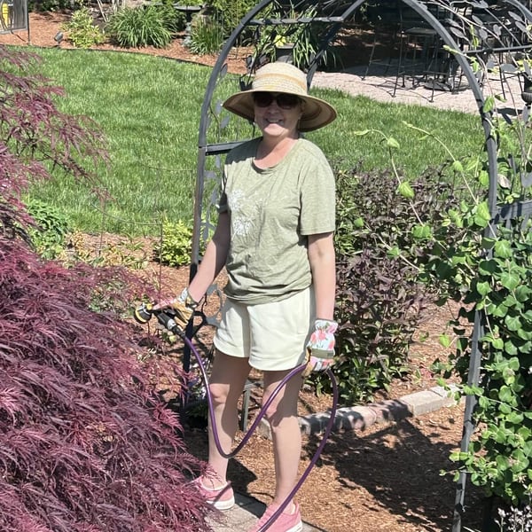 Holly Folgia smiling in a sunny backyard garden while holding a watering hose.