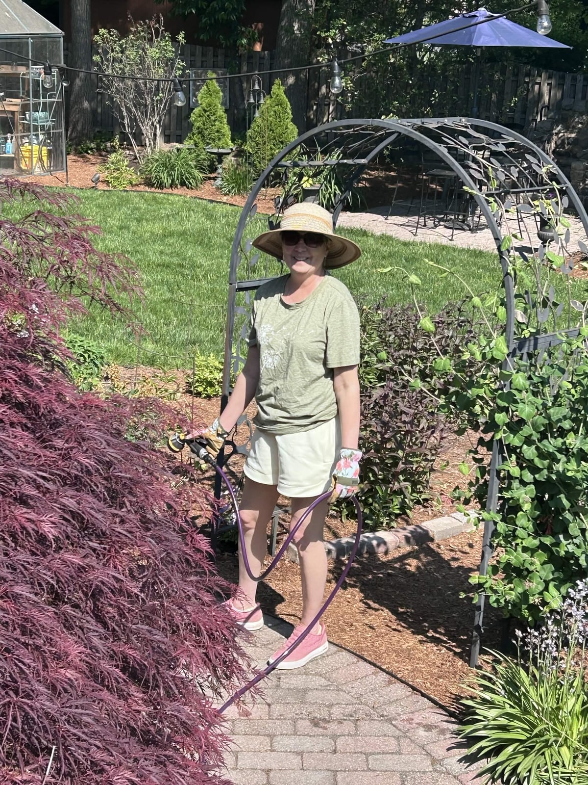 Holly Folgia smiling in a sunny backyard garden while holding a watering hose.