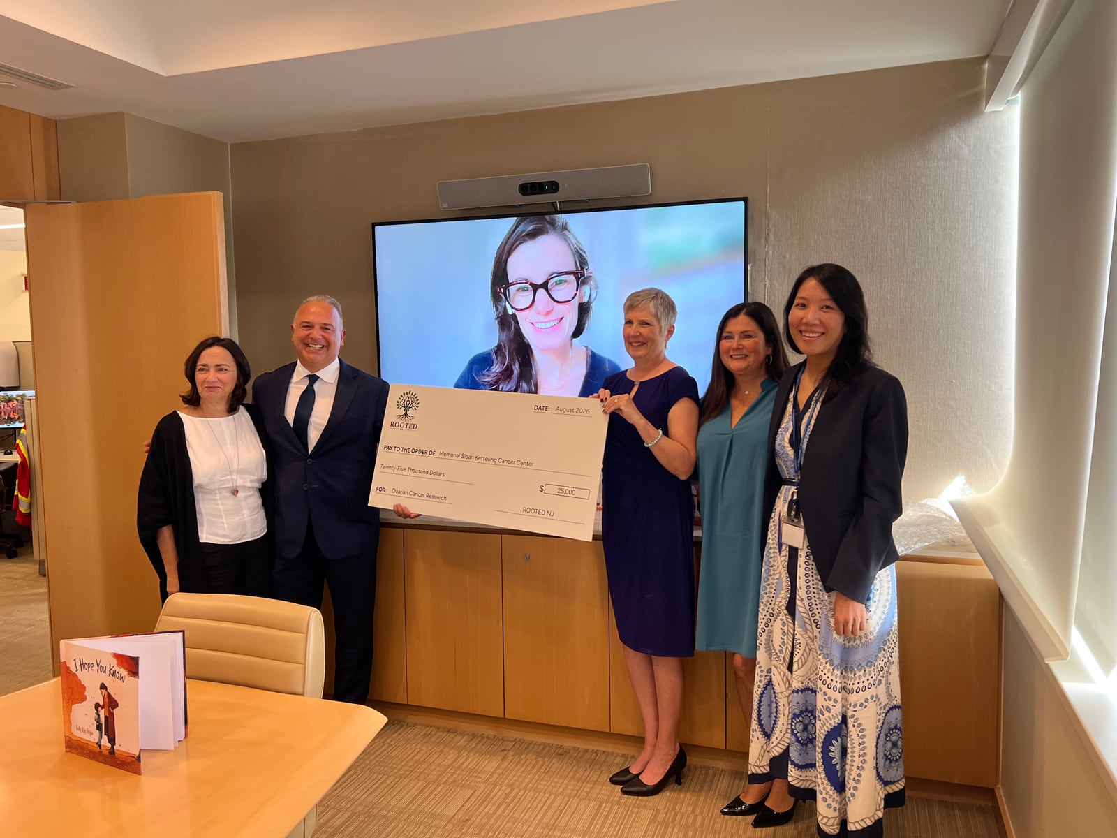 Attendees holding a $25,000 ceremonial check beneath a video-call display, with I Hope You Know on the table.